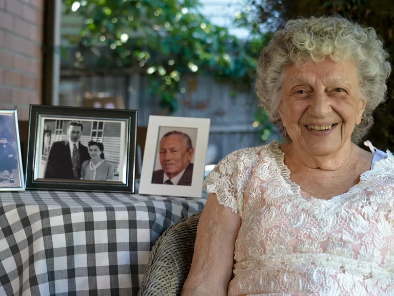 Smiling woman wearing a pink and lace top, sits beside a table with 3 framed photos from her life.