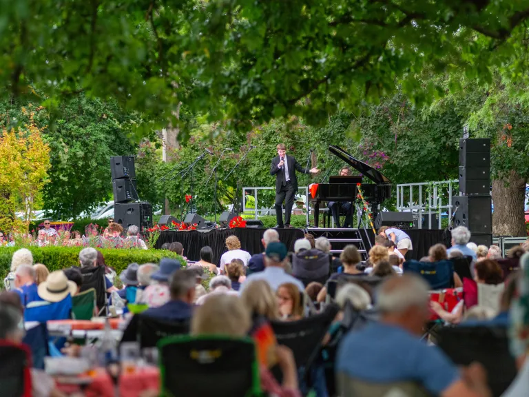 A performer and pianist on stage in front of a seated audience in a park
