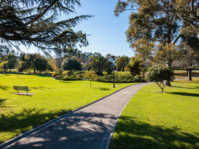 A path cuts through lush green grass leading toward neatly manicured gardens. Tall trees lean in from the sides, shading sections of the path.