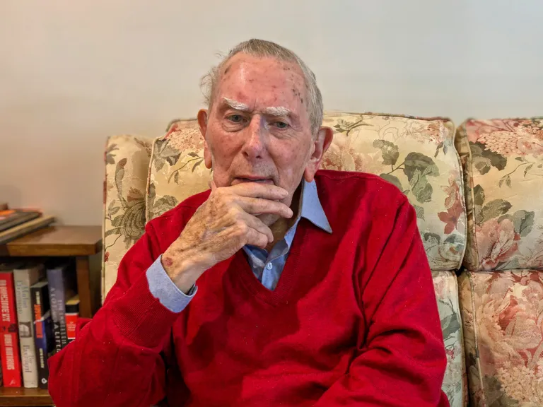 Man sitting on a floral couch. Lookslike he is pondering something. He's wearing a blue collared shirt under a red jumper.
