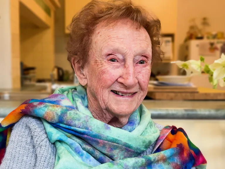 Smiling woman with short auburn hair, sits at the table with the kitchen behind her. She's wearing a scarf that is mostly shades of blue, but has bright pops of red and orange.  