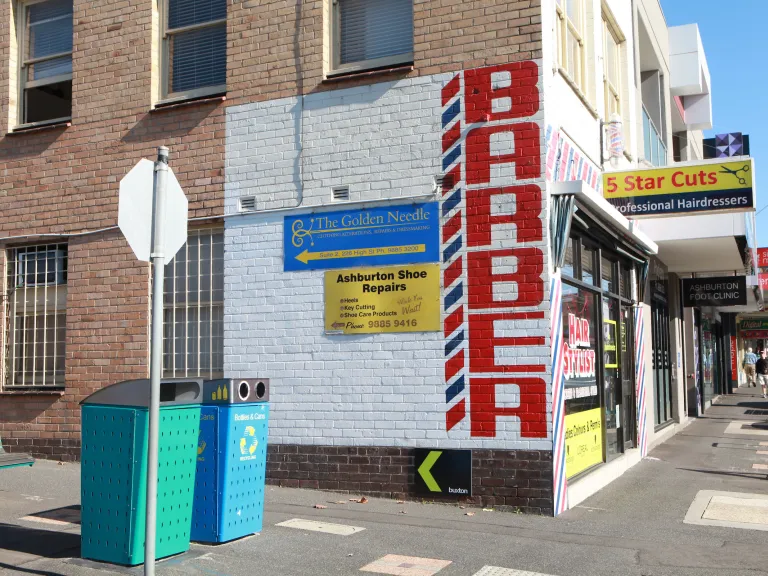 A rubbish and recycling bin on a street corner next to a barber shop.