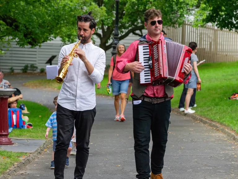 An accordion player and oboe player casually walk along a path playing their instruments.