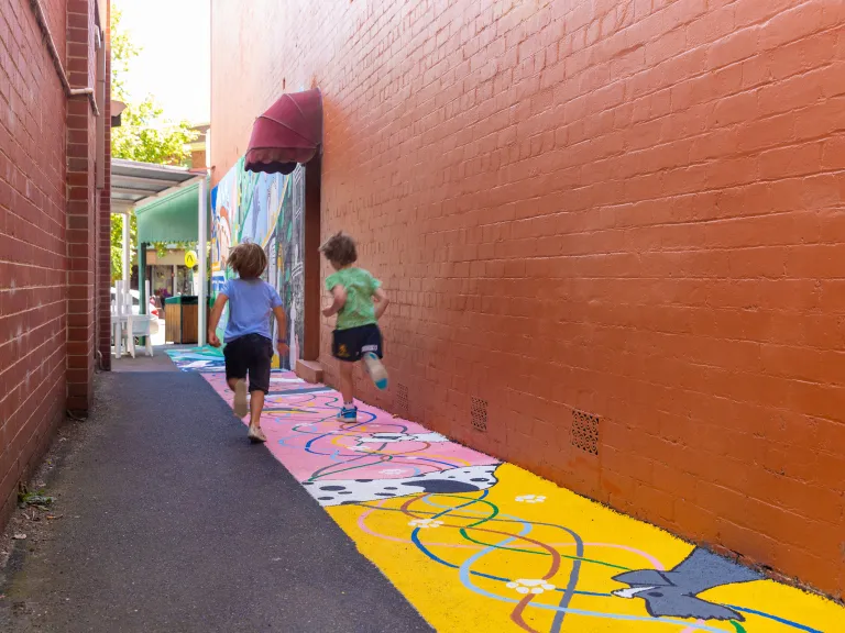 Two children run down a laneway with red/brown painted wall and a dog mural on the ground.
