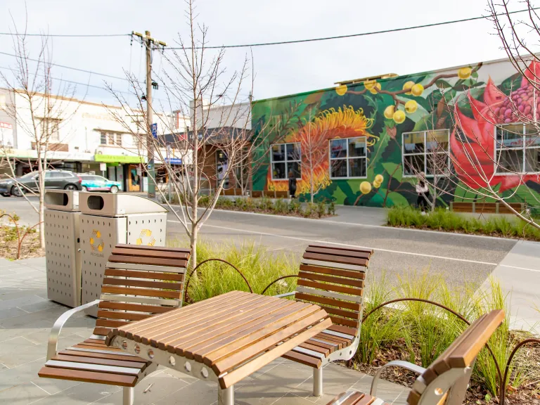 A wooden topped table and seats looking fresh. They're in a clean streetscape with plants and bins.