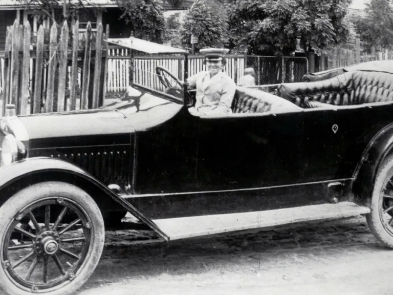 Black and white photograph of a women in an old-fashioned car with no roof. The women is wearing a jacket and cap and smiling into the camera.