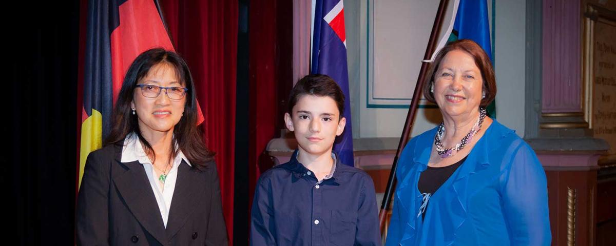 Dr Elaine Ong, Aidan Dimitriadis and Yvonne Giltinan stand on the podium with flags behind them at the Australia Day ceremony