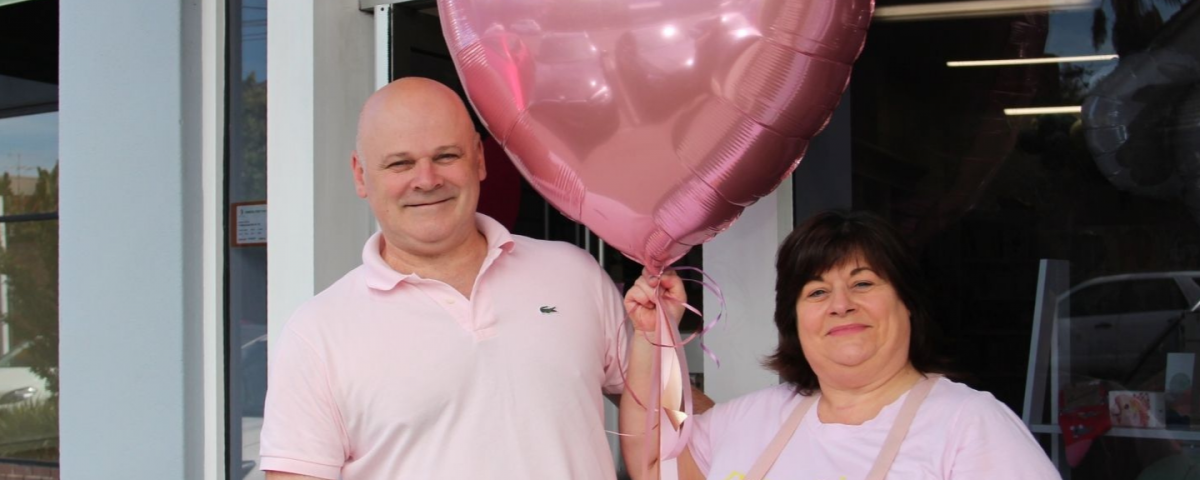 Two business owners stand outside of their business holding a giant pink balloon