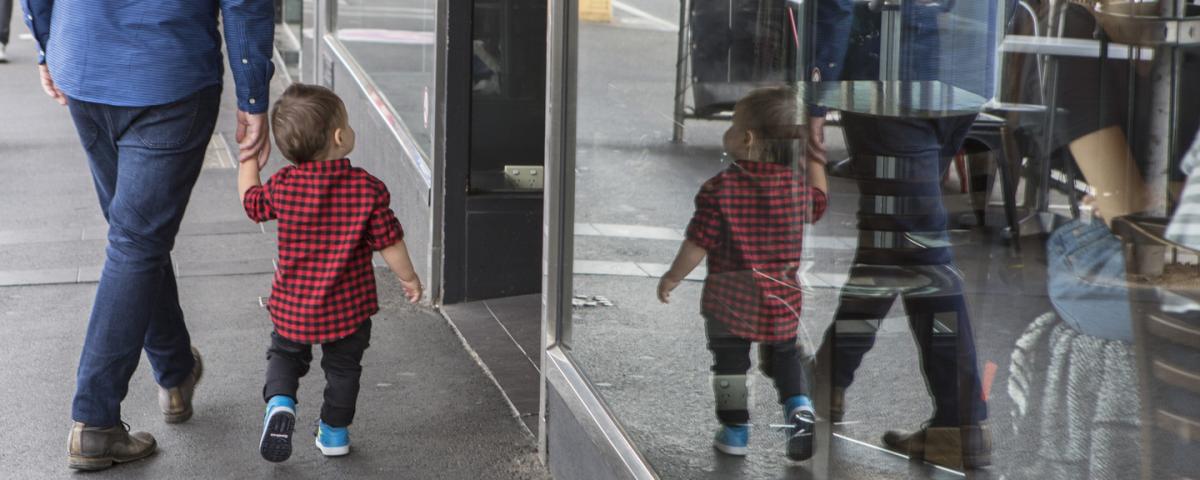 An adult and a child are walking down a street their reflectons are shown in the window of a restaurant.