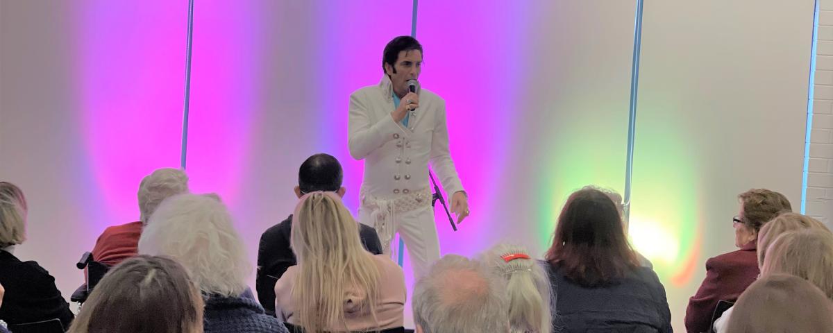An Elvis tribute performer singing in a white jumpsuit in front of a seated crowd