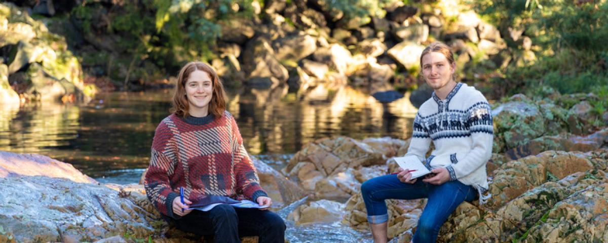 A man and woman sit on rocks at a creek with their feet in the water