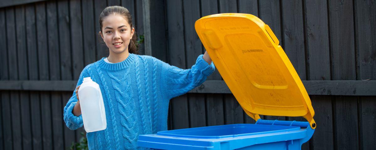 A young person holding an empty milk carton next to an open recycling bin