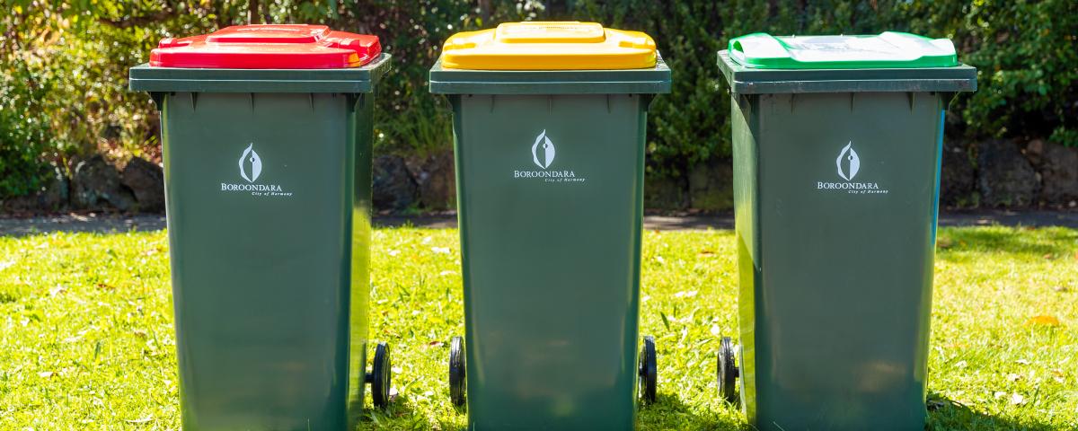 3 green bins on a lawn, one with a red lid, one with a yellow lid and one with a light green lid