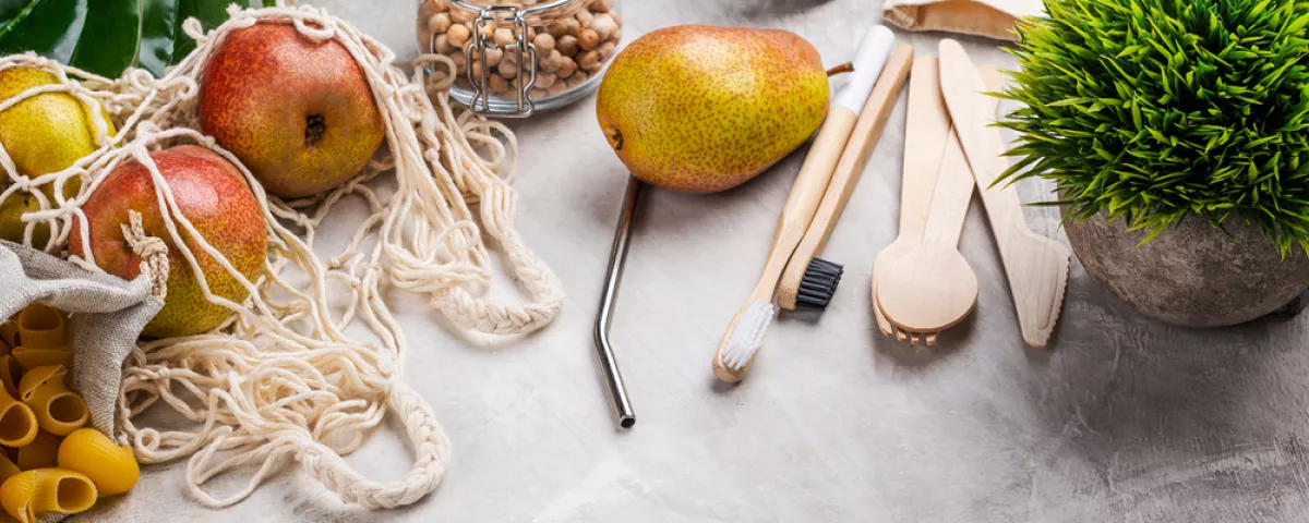 A close-up of a table with pasta, fruit, and nuts all in sustainable containers or bags, and wooden cutlery and toothbrushes with a metal straw and a plant alongside