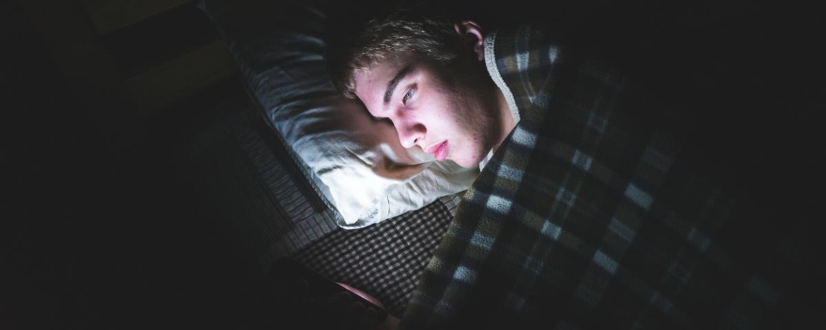 A young person lying in bed in darkness with their face brightly lit up by a mobile phone screen that they are using