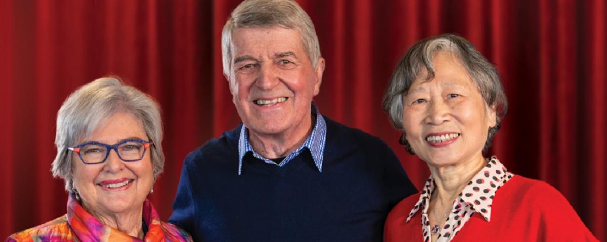 3 older people smile in front of a red velvet curtain