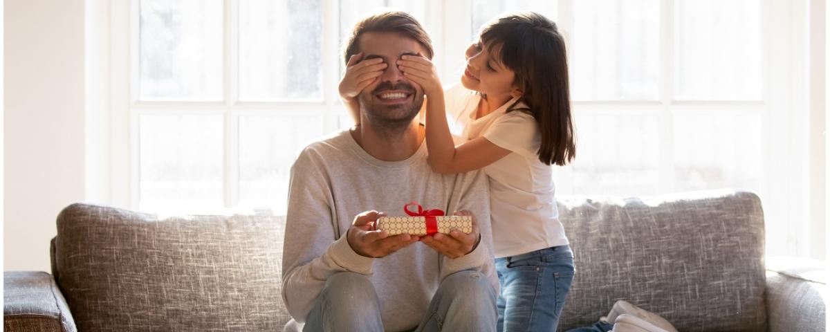 A young girl covers her father's eyes while he holds an unwrapped gift and smiles
