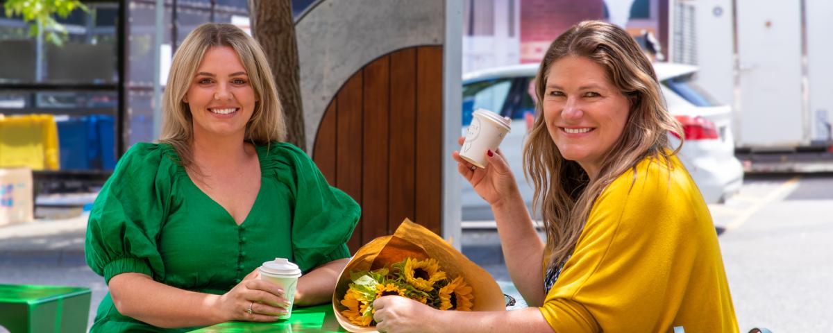 2 brightly-dressed people smile as they sip coffee at a green table