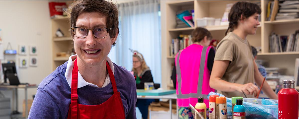 Artist wearing purple top and red apron smiles in studio.