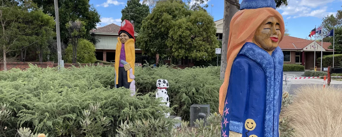 A garden bed with low shrubs and 3 wooded carved sculptures. 1 of a dalmation, and 2 people, one a fireman and the other a woman wearing a long blue coat and matching hat.