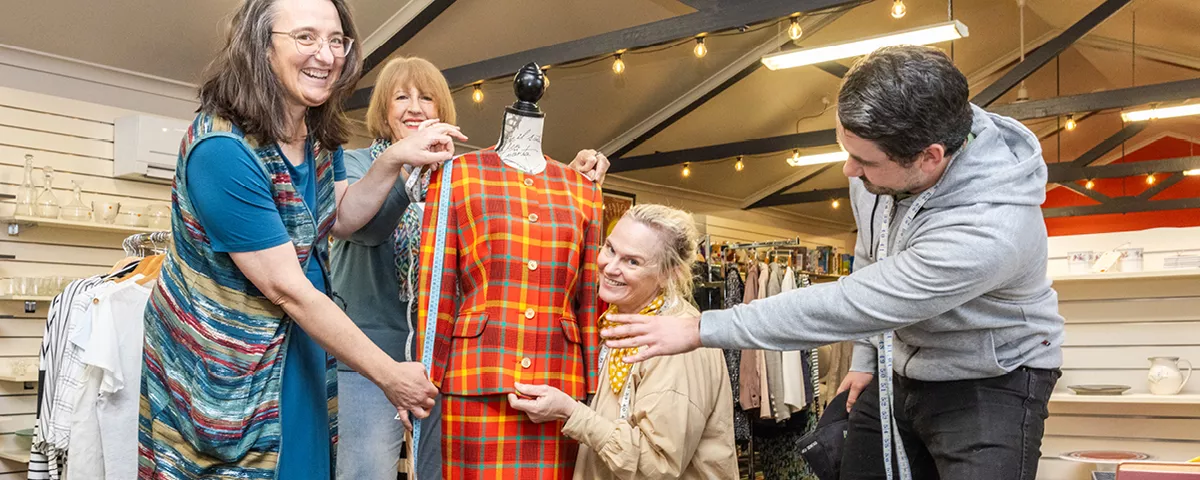 Four op shop volunteers holding measuring tapes stand with dressmaker dummy wearing brightly coloured checked suit.