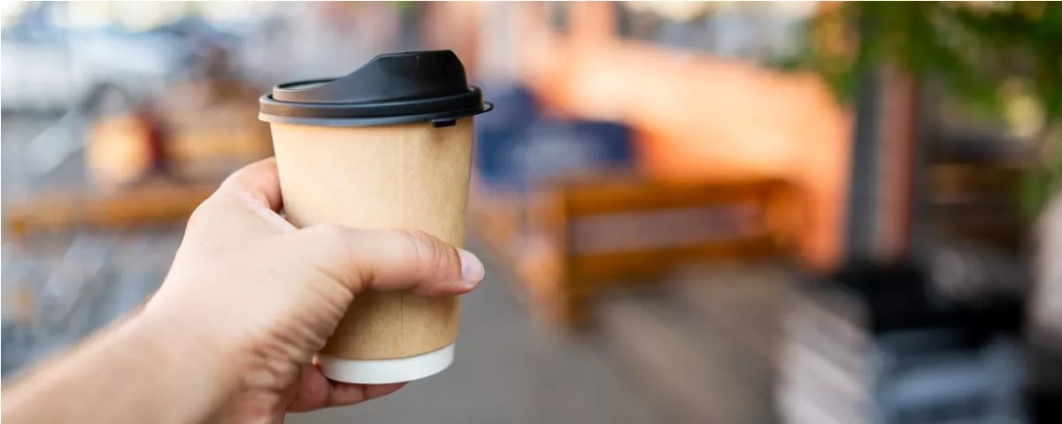 Person's hand holds disposable paper coffee cup with plastic lid
