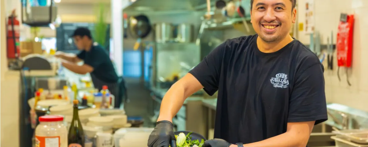Man stands smiling in café kitchen while emptying vegetable scraps into container.