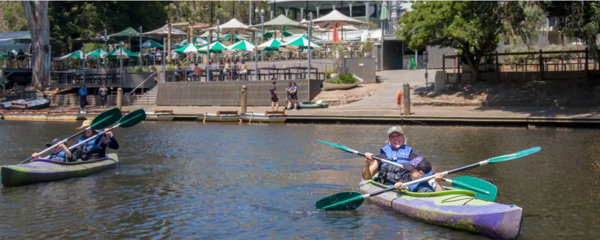 Woman and child wearing life vests in kayak on river outside building with café umbrellas.