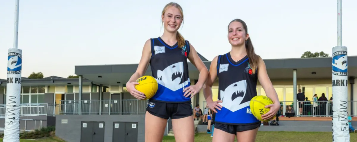 Two teenage girls in blue and black football uniforms stand smiling outside pavilion.