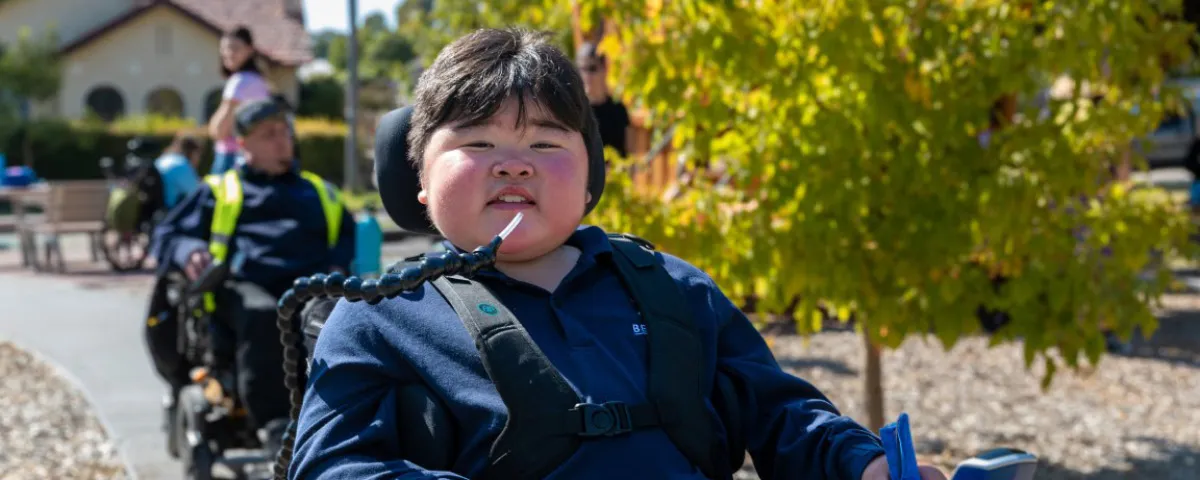 Three people in wheelchairs on path in playground, with boy smiling at front and house and trees in background.