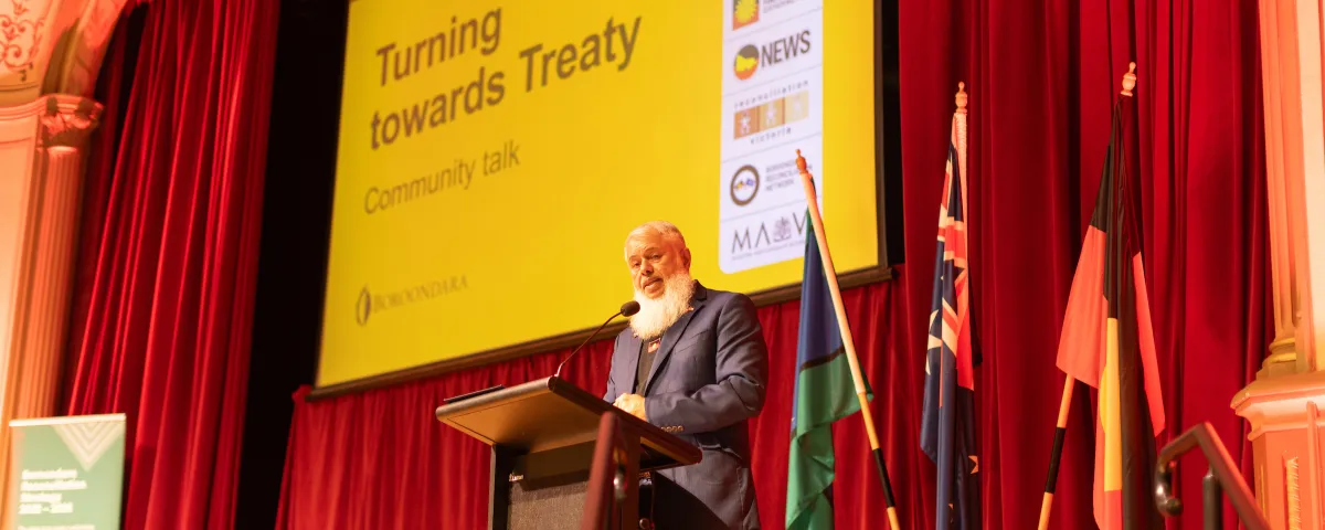 A man stands on stage behind a lectern, with a PowerPoint presentation displayed in the background.