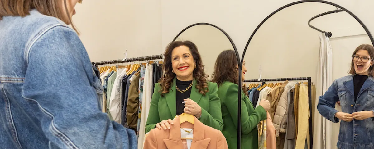 2 women in a clothing store, one is the sales assistant holding up a jacket while the other is a customer trying on a jacket.