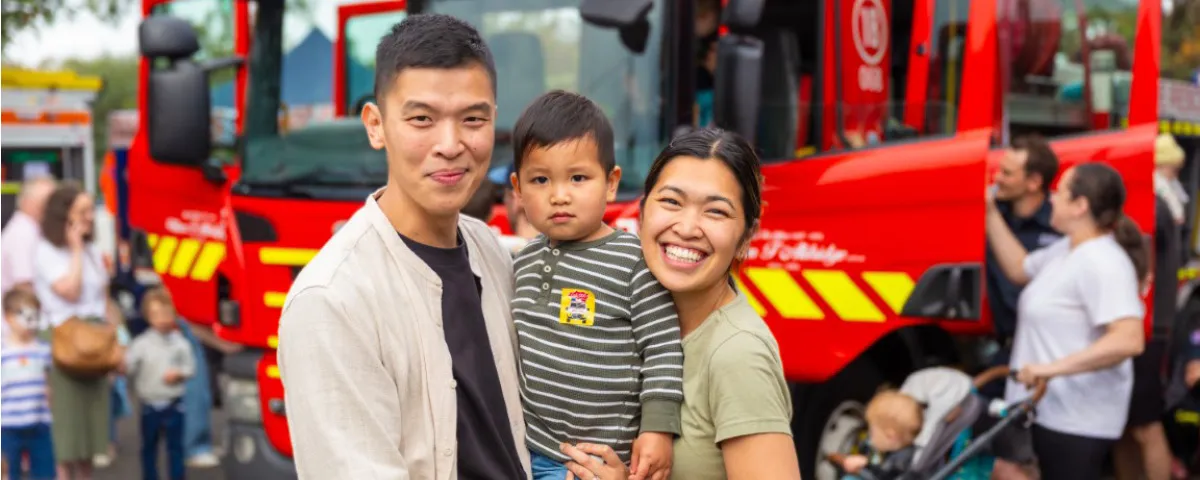 Smiling man and woman stand holding small child with fire engine in background