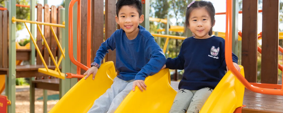 Two children sit on two slides at a playground.