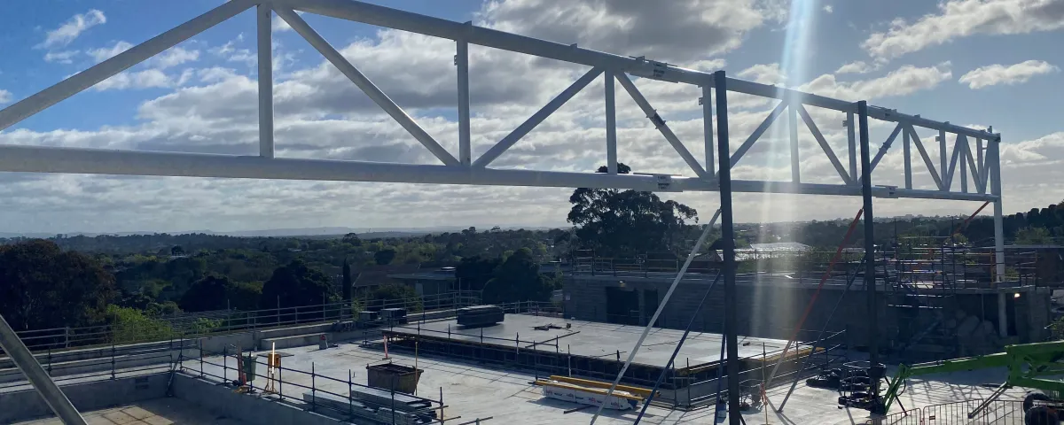 A construction site under a bright sky with scattered clouds. A large metal beam structure is in the foreground, and unfinished concrete structures, safety rails, and equipment are visible in the midground. Distant trees and hills complete the background.