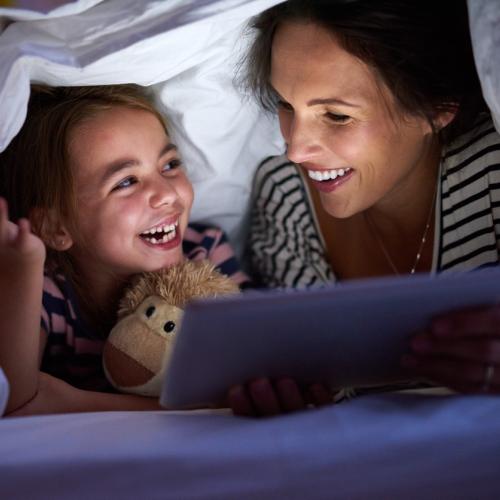 A young girl and her mother, with a bed sheet over their heads, as they read by torchlight