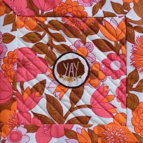 A brightly coloured quilt with flowers and a patch in the middle that says 'YAY' and is decorated with flowers
