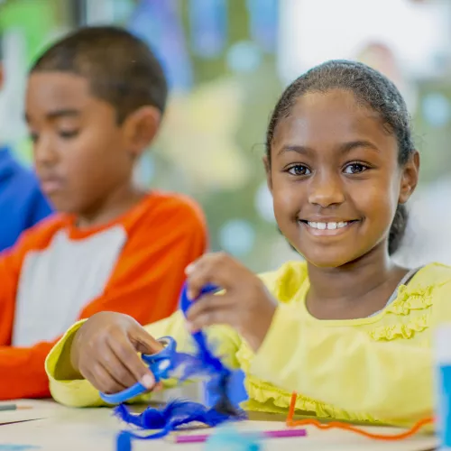Three children participating in craft. Child in yellow shirt in focus, holding a pair of scissors cutting a feather.