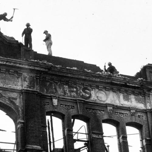 A black and white photo of people working on top of an old building facade which is damaged and burnt