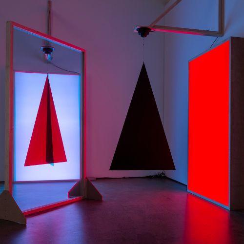 A mirror reflecting the image of a red triagle with a black line through the centre, next to a bright red lit rectange and a hanging 3D black pyramid in front of it.