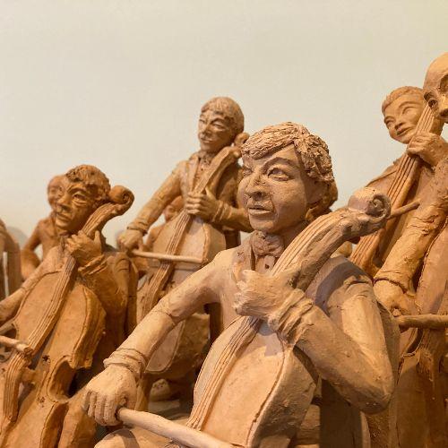 Unpainted clay sculptures of five figures playing cellos. Three of the figures are seated while the remaining two are standing up, behind them. In the background is another clay figure, standing and blowing a horn and another seated figure whose instrument is obstructed from view. 