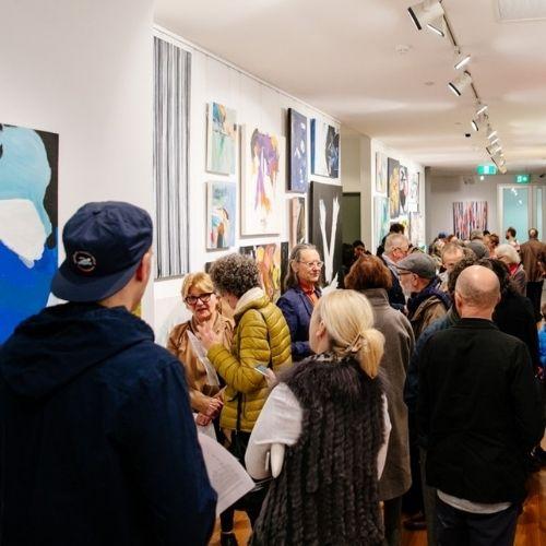 Crowd of people look at paintings hanging in a narrow gallery space.