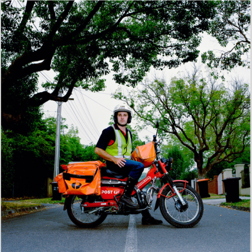 Photograph of a postman on a red Australia Post motorcycle. Orange mailbags are attached to the bike, and the postman is wearing a high-vis vest and white helmet. He is stopped in the middle of the street with one foot on the ground to steady the bike looking directly at the camera. The background shows a tree-lined street with wheely bins out for collection.