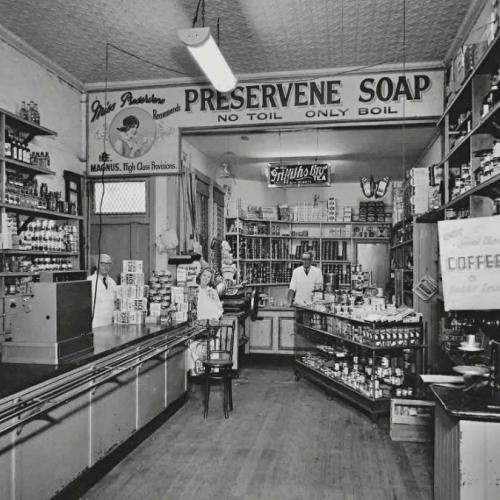 Black and white photograph of a grocer shop. Shelves lined with tinned and bottled goods line the walls of the store and there is a long table with a cash register. Three staff are in the background, one behind a counter at the back of the shop and two near the register partially concealed by product displays.