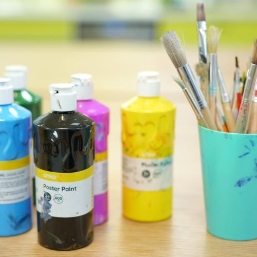 Bright coloured paint bottles and a cup filled with used paint brushes on a table. 