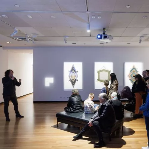 Image of man in gallery speaking to a crowd of people. Artwork is hung on wall in background. 