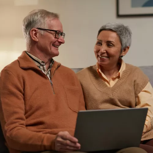 A man and woman sitting on a couch looking at each other smiling. The man has a laptop on his lap.