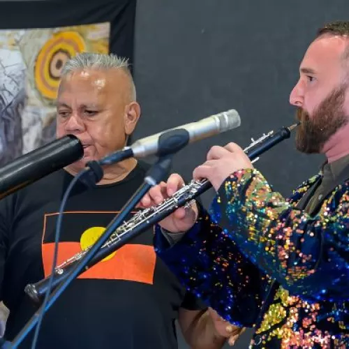 Three people performing. A man in a sparkly blue jacket plays the flute. Another man in a black t-shirt featuring the Aboriginal flag is playing the didgeridoo and a woman in the background plays cello