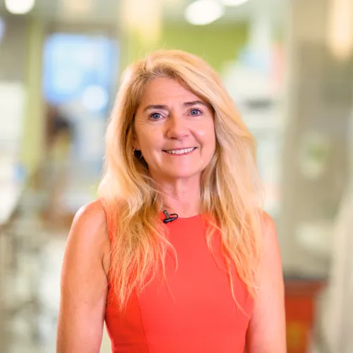 A woman with long blond hair is looking at the camera smiling. She is wearing an orange sleeveless top.