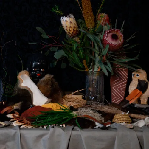 A vase with flowers sits on a grey tablecloth. In front of the vase is a cluster of plants, shells, reed baskets and dolls. 